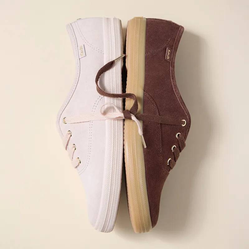 The Keds Champion Luxe Suede Lace-Up Sneaker in blush and the Keds Champion Luxe Suede Lace-Up Sneaker in chocolate, both on their side, soles together with the laces tied together artfully. The golden eyelets, aglets, and Keds logo are clearly visible.