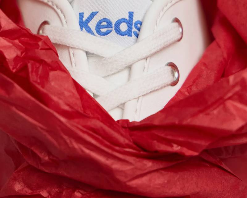A close-up of the Keds Champion Originals Lace-Up in white wrapped inside red tissue paper. The text says Perfect Gift, Perfect Fit. Explore Holiday Gift Shop.