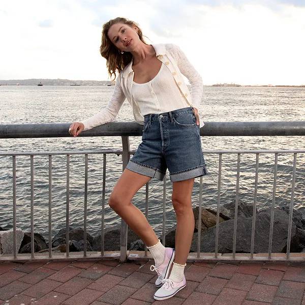 A woman wearing cuffed denim shorts, a white top and open cardigan, and the Keds Champion sneakers in colorblocked pastel canvas with a seascape horizon in the background.